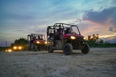 Red side-by-side UTVs driving on a dusty dirt trail at dusk, headlights glowing as helmeted riders enjoy a sunset off-road adventure.