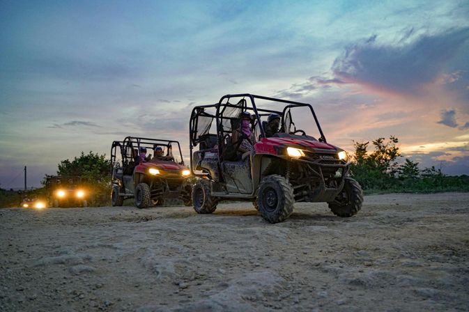 Red side-by-side UTVs driving on a dusty dirt trail at dusk, headlights glowing as helmeted riders enjoy a sunset off-road adventure.