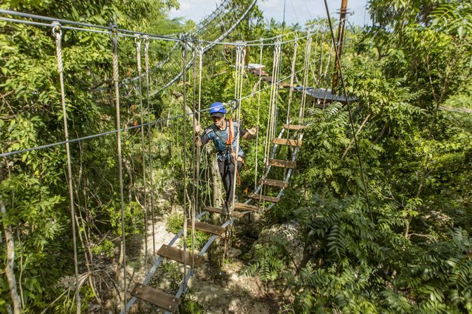 Person in helmet and harness crossing a suspended wooden-plank rope bridge through a lush tropical rainforest canopy on an outdoor aerial adventure course.