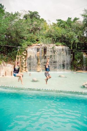 Two people in life vests ziplining over a turquoise lagoon toward a cascading rocky waterfall set in lush tropical jungle.