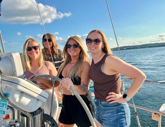 Four smiling friends in sunglasses steering a sailboat on a sunny lake with blue sky, gentle waves, and a distant shoreline — summer sailing fun.