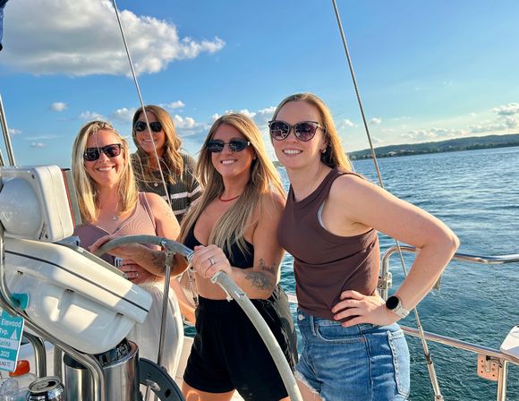 Four smiling friends in sunglasses steering a sailboat on a sunny lake with blue sky, gentle waves, and a distant shoreline — summer sailing fun.