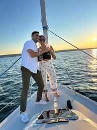 Smiling couple toasting with champagne on a sailboat at sunset, golden sun reflecting on calm coastal waters