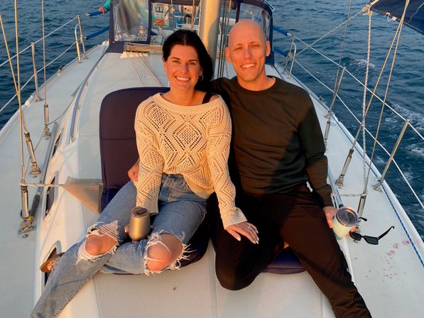Smiling couple relaxing on the bow of a sailboat during golden hour, holding travel mugs with ocean, rigging and sunset light in the background