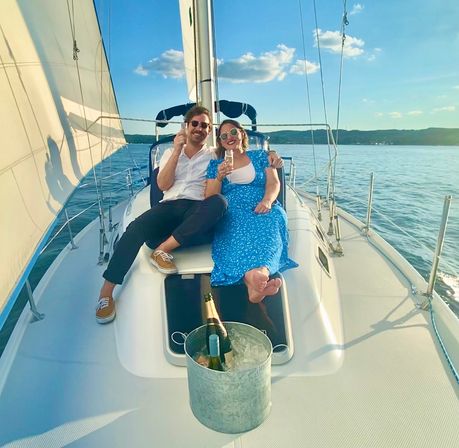 Two people relaxing on the bow of a sailboat on calm blue water near a distant shoreline, sipping champagne with an ice bucket in the foreground and white sails under a sunny sky.