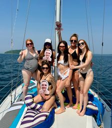 Seven women in swimsuits smiling and posing with cardboard face cutouts on a sailboat, blue lake and tree-lined shoreline under a clear summer sky.