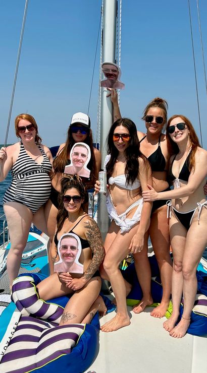 Seven women in swimsuits smiling and posing with cardboard face cutouts on a sailboat, blue lake and tree-lined shoreline under a clear summer sky.