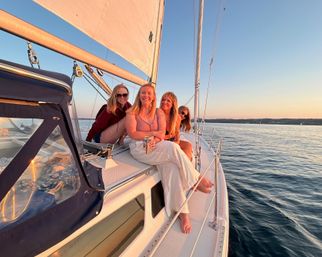 Four smiling friends sitting on a sailboat deck at golden sunset, casual summer outfits and a raised sail overhead, cruising on calm blue water with a distant shoreline.