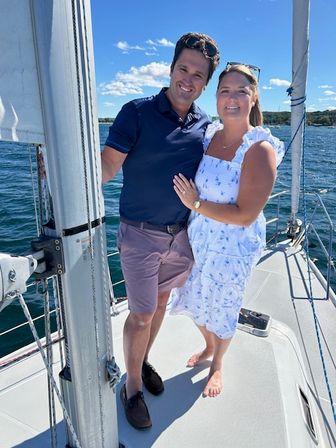 Smiling couple posing on a sailboat deck during a sunny summer sail — woman in a white floral sundress barefoot and man in a navy polo and shorts with blue sky and sparkling water behind them.