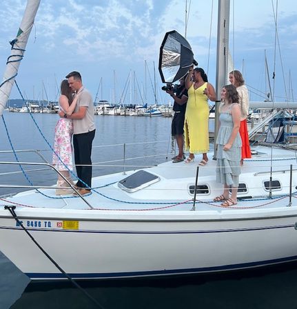 Couple embracing on the bow of a sailboat during a romantic photoshoot at a marina at dusk, photographer with a softbox light and three attendants standing on deck, rows of moored boats and calm harbor water in the background.