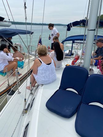 Group of people relaxing on a sailboat with blue cushions, drinks and snacks on a calm lake near a tree-lined shore under an overcast sky.
