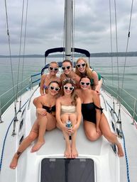 Six friends in swimsuits and heart-shaped sunglasses pose on the bow of a sailboat on a calm lake; bride-to-be in white sits center holding a drink.