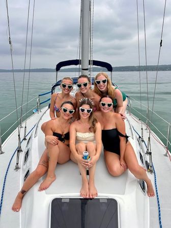 Six friends in swimsuits and heart-shaped sunglasses pose on the bow of a sailboat on a calm lake; bride-to-be in white sits center holding a drink.