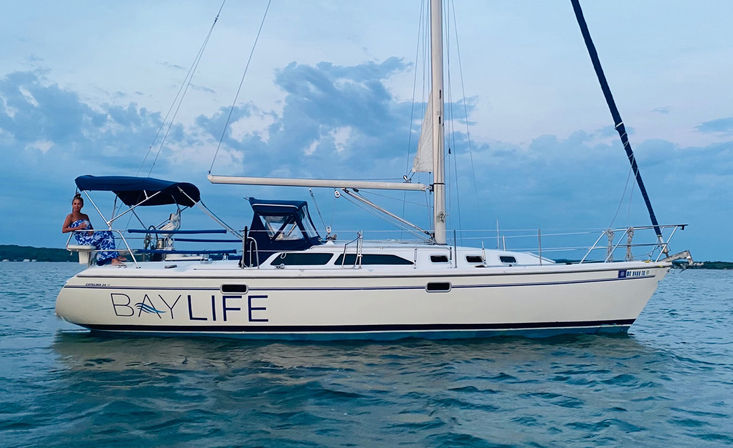 White sailboat with blue canvas bimini anchored on calm coastal waters at twilight, tall mast and cloudy sky, person relaxing on the stern