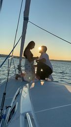 Romantic sailboat sunset on calm coastal waters — one person kneeling on the bow as their partner stands, sun glowing between them.