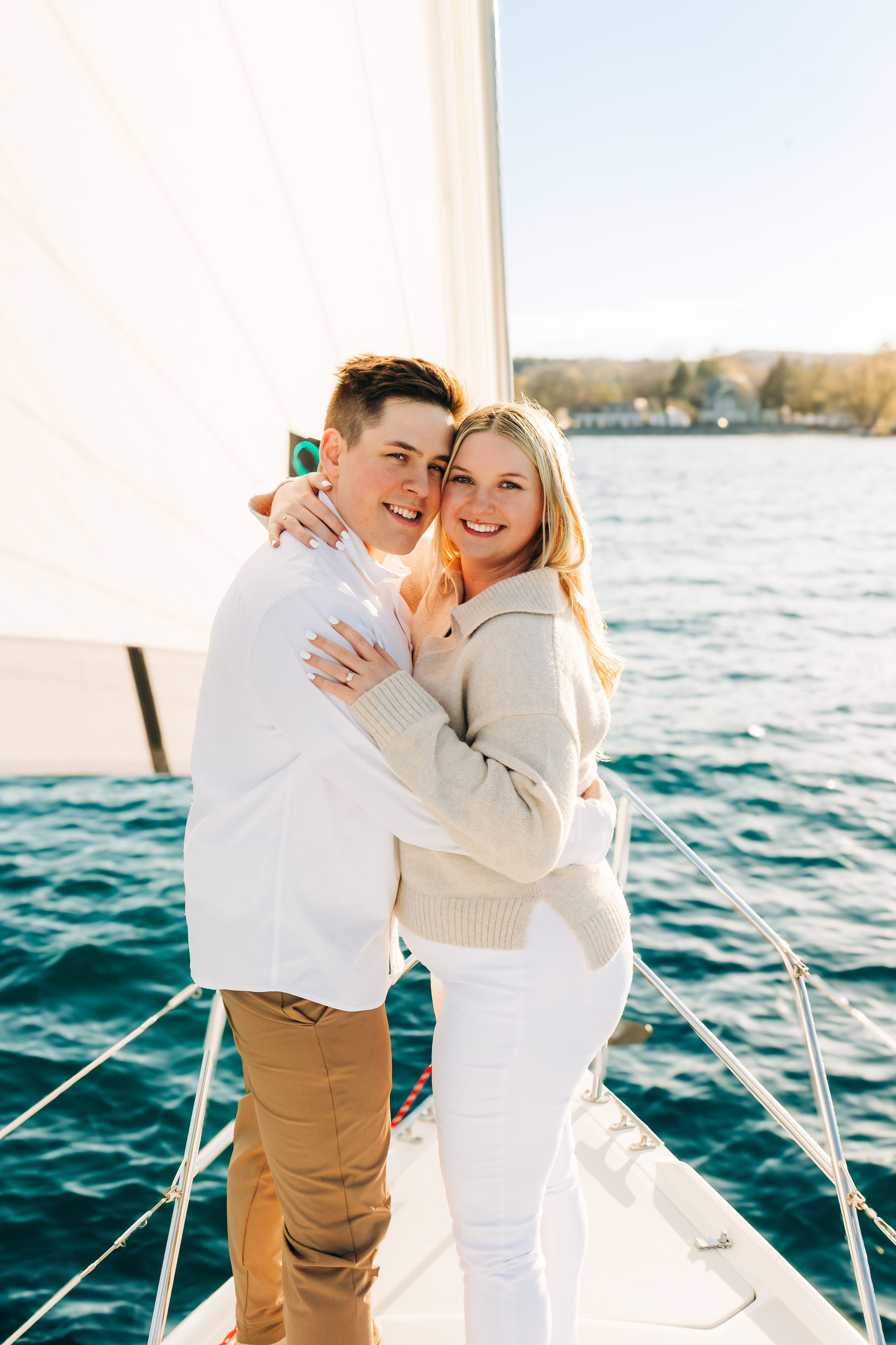 Beaming couple embracing on the bow of a sailboat, white sail and blue water behind them with a sunlit shoreline in the distance.