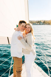 Beaming couple embracing on the bow of a sailboat, white sail and blue water behind them with a sunlit shoreline in the distance.