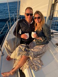 Smiling couple in sunglasses holding insulated mugs while sailing at sunset, wrapped in patterned blankets on a sailboat with white sails and calm blue ocean in the background.