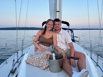 Smiling couple seated on the bow of a sailboat at dusk on calm lake waters, with a metal ice bucket and champagne between them.