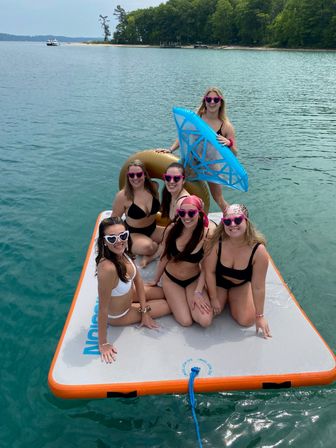 Six friends in bikinis wearing heart-shaped sunglasses smiling on an inflatable lake platform, holding a gold donut and blue float with clear turquoise water and a tree-lined shoreline in the background — summer lake day