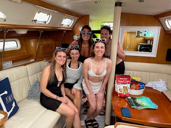 Five friends in swimsuits and sunglasses smiling inside a sailboat cabin, seated on cream leather benches around a wooden table with snacks and magazines — cozy yacht interior scene.