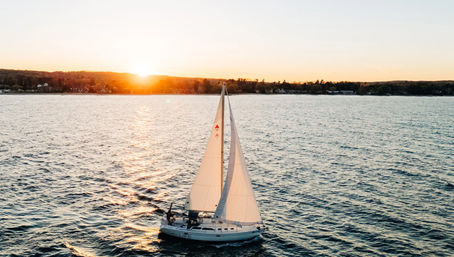 Sailboat gliding across open water at sunset, white sails catching golden light with a tree-lined shoreline and low hills on the horizon.