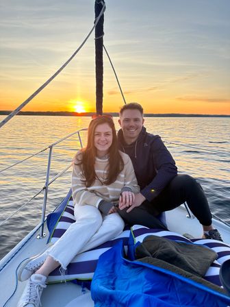 Smiling couple holding hands on the bow of a sailboat during a golden sunset over a calm lake.