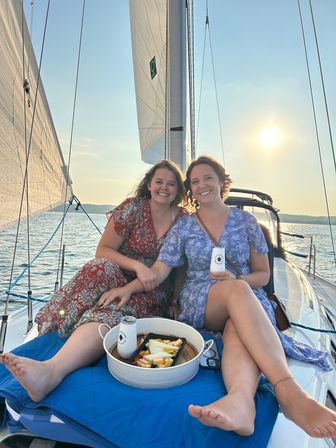 Two smiling friends relaxing on a sailboat at sunset over a calm lake, sharing a cheese-and-fruit picnic.