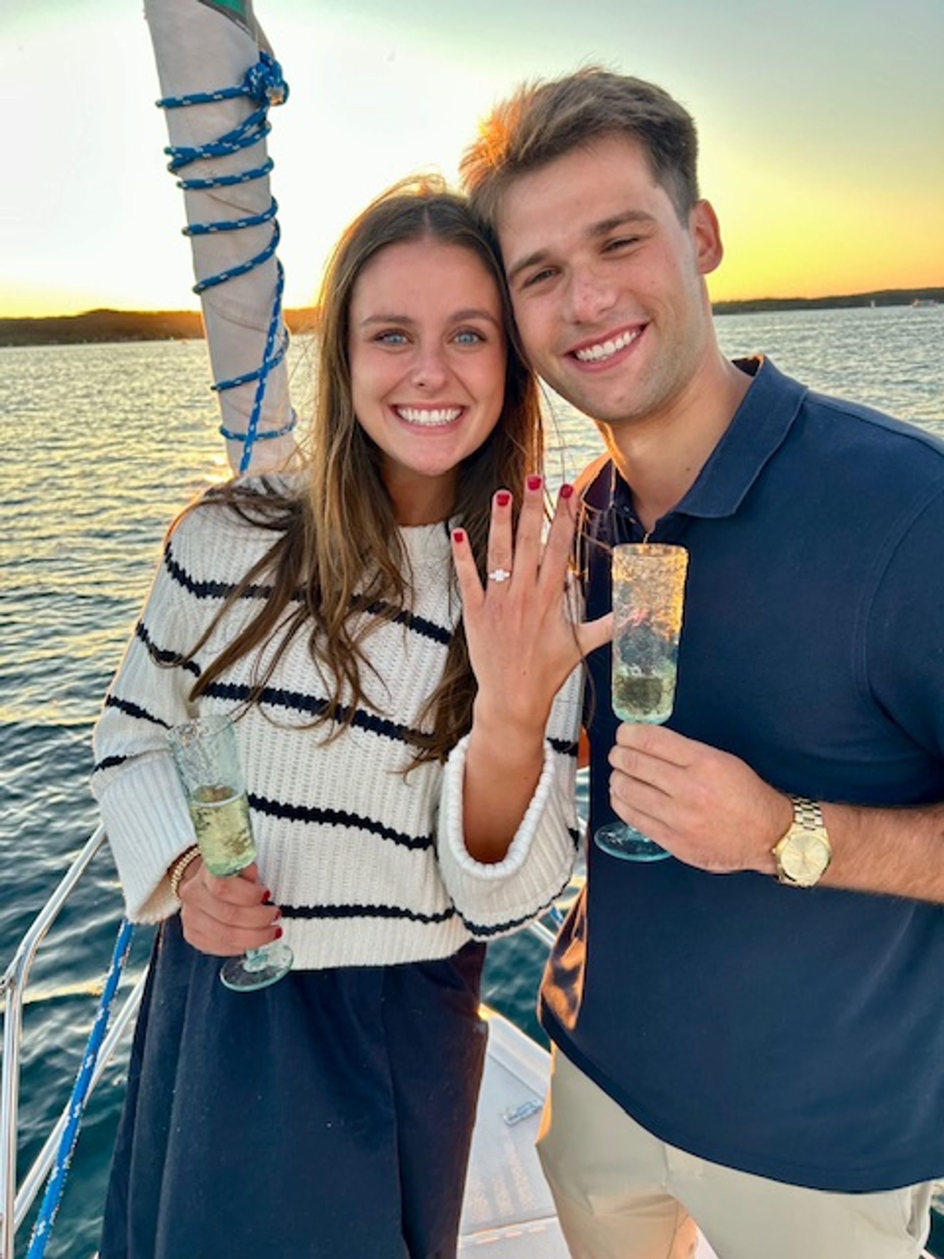 Young couple on a sailboat at sunset, smiling and holding champagne flutes as the woman shows off her engagement ring over calm open water