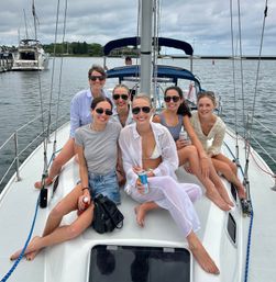 Group of friends smiling on a sailboat deck at a marina harbor, wearing summer outfits and sunglasses and enjoying drinks under a cloudy sky