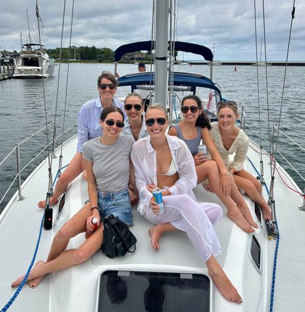 Group of friends smiling on a sailboat deck at a marina harbor, wearing summer outfits and sunglasses and enjoying drinks under a cloudy sky