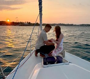 Sunset sailboat proposal: a person kneels on the bow holding their partner's hand on a sailboat with a marina and setting sun over rippling water