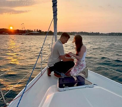 Sunset sailboat proposal: a person kneels on the bow holding their partner's hand on a sailboat with a marina and setting sun over rippling water