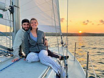 Smiling couple on the bow of a sailboat during a golden sunset, white sails and rigging, rippling water and distant shoreline