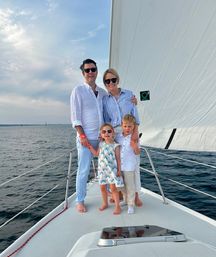 Smiling family of four on the bow of a sailboat—parents and two kids in summer clothes and sunglasses, large white sail and calm open water under a partly cloudy sky.