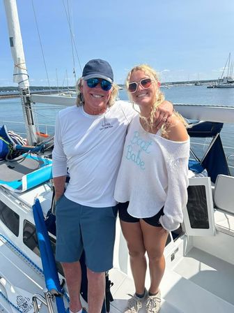 Two smiling people — a man in a cap and captain-style shirt and a woman in a 'Boat Babe' sweater — posing on a sunny sailboat deck with a marina and calm water in the background.
