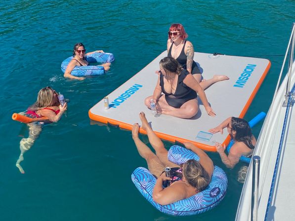 Sunny boat party with a group relaxing on a large inflatable floating mat and blue patterned inner tubes in clear teal water.