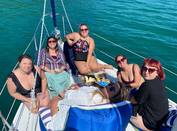 Six friends relaxing on the bow of a sailboat, smiling in sunglasses and swimsuits and sharing picnic snacks under bright sun on clear turquoise water.
