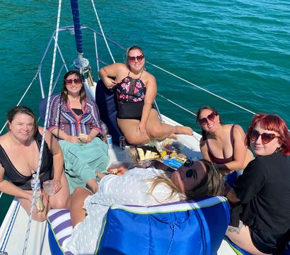 Six friends relaxing on the bow of a sailboat, smiling in sunglasses and swimsuits and sharing picnic snacks under bright sun on clear turquoise water.