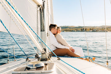 Couple cuddling on a sailboat deck with white sails and blue rigging, cruising a calm lake past a tree-lined shoreline on a sunny day.
