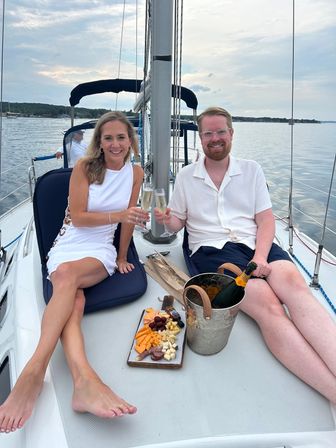 Smiling couple clinking champagne glasses aboard a sailboat with a charcuterie board and ice bucket on the deck, calm lake and tree-lined shore in the background.
