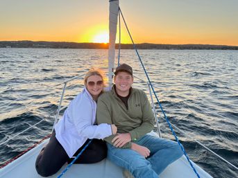 Smiling couple cuddled at the bow of a sailboat on a sunset sail, rippling lake waters and distant shoreline bathed in golden light.