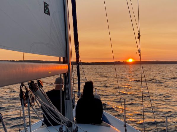 Silhouetted people on a sailboat bow framed by mast and rigging, watching a golden-orange sunset over rippling coastal waters