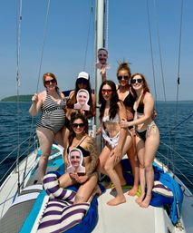 Seven friends in swimsuits posing on a sailboat deck holding printed face cutouts, sunny blue sky and calm open water with a tree-covered island in the distance — summer boating outing.
