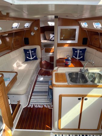 Bright sailboat cabin interior with teak floors and wood trim, cream leather seating, compact galley sink, striped rug and navy anchor throw pillows saying Sail Away.