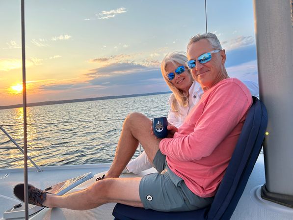 Couple wearing mirrored blue sunglasses enjoying a sunset sail on calm water, seated on a sailboat deck holding a navy anchor cup with golden sky reflecting on the waves
