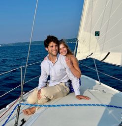 Smiling young couple lounging on the bow of a sailboat, barefoot and relaxed, sailing on a deep-blue lake under a clear sky.