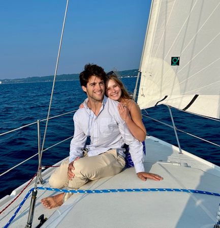 Smiling young couple lounging on the bow of a sailboat, barefoot and relaxed, sailing on a deep-blue lake under a clear sky.