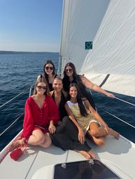 Five women relaxing on the bow of a sailboat on a sunny summer day, wearing sunglasses and summer dresses, with a large white sail and blue open water in the background.