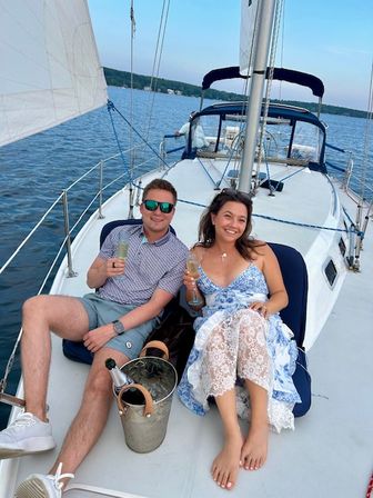 Smiling couple relaxing on a sailboat deck, toasting with champagne flutes and an ice bucket, enjoying a sunny summer day on a calm lake with a tree-lined shoreline.
